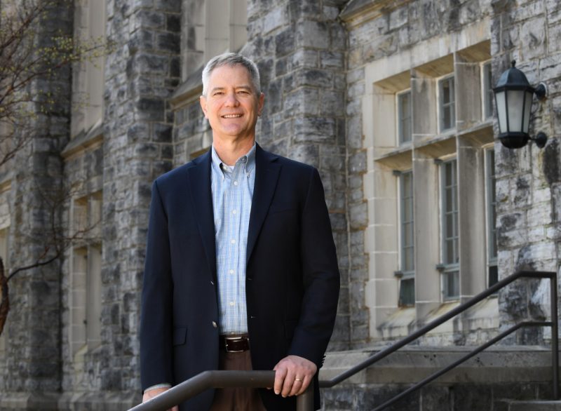 Wearing a blue sports coat and dress shirt, Kevin Pitts stands on some stairs, with Hokie Stone building behind him.