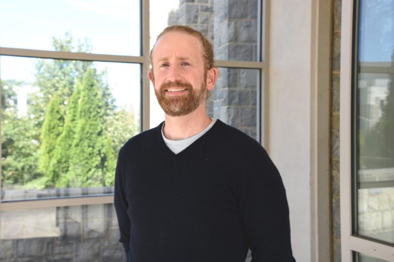 A man wearing a v-neck sweater poses for the camera outside a lab building.