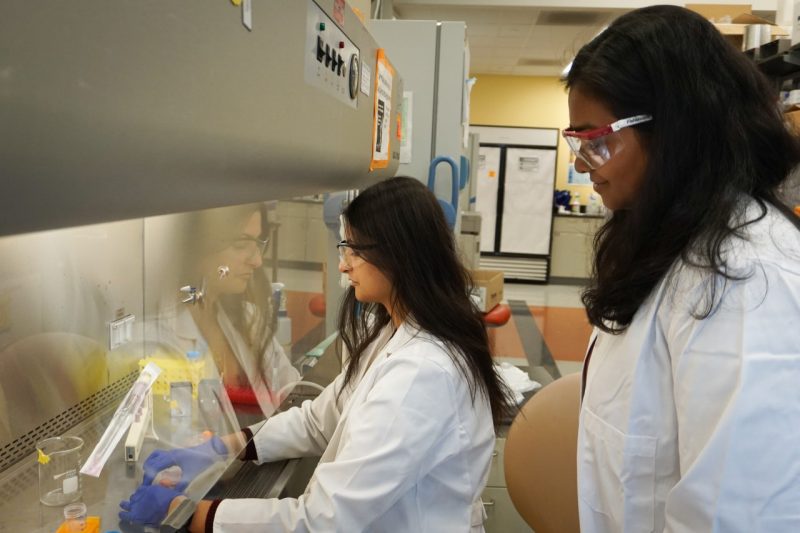 Two women wearing white lab coats and safety glasses look into a laboratory cabinet, where one of them is holding small vials. 