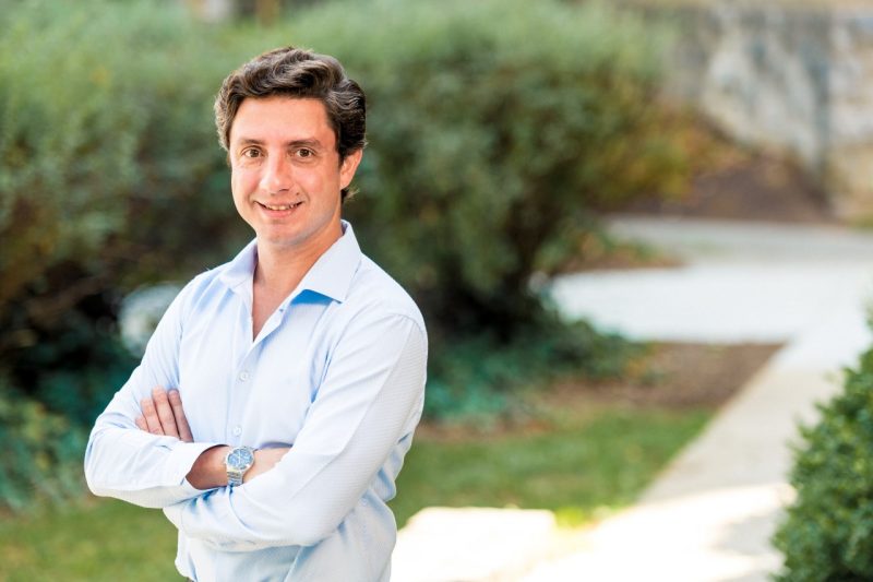 A man in a blue dress shirt poses with his arms folded on a sidewalk.