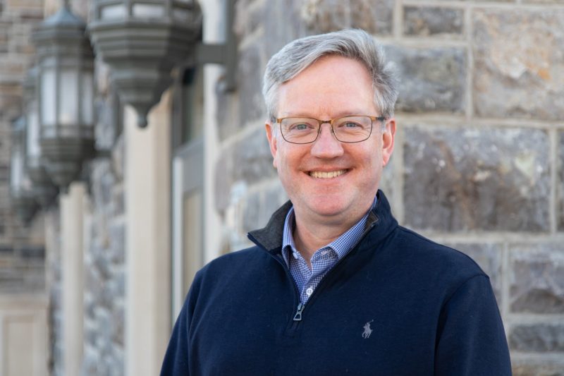 A man with salt and pepper hair, eyeglasses, and a blue sweater over a dress shirt smiles as he stands near Davidson Hall.