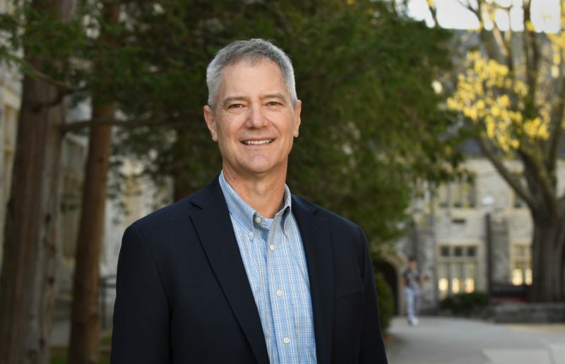 A man with salt and pepper hair stands on a campus sidewalk,, wearing a dress shirt and sports coat.