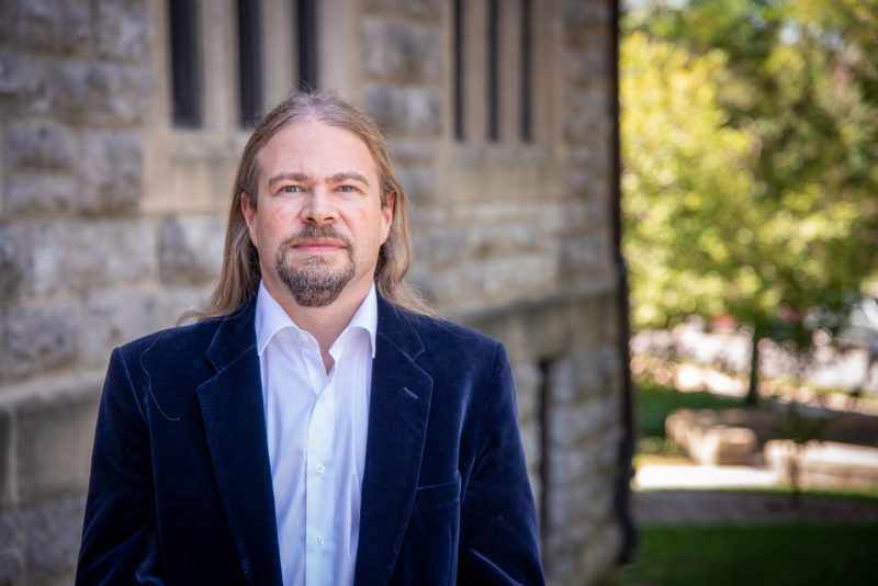 A man with long black brown hair and a beard stands by a Hokie Stone building, wearing a blue sports jacket over a blue dress shirt.