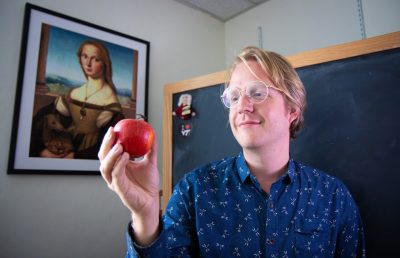 Dan Hoek holds an apple in front of a chalkboard and a portrait
