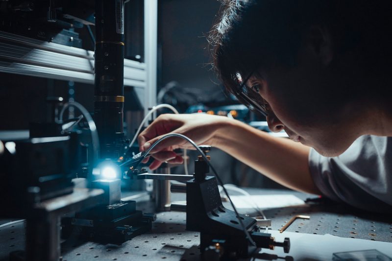 A researcher leans over a soldering tool, which is sparking blue. Dramatic lighting.