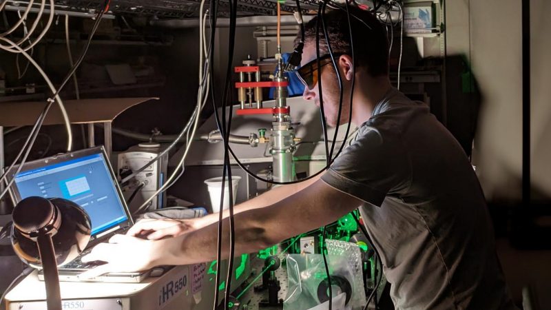 Yannick Pleimling, wearing protective eyegear, works on a computer in a dark lab filled with equipment.